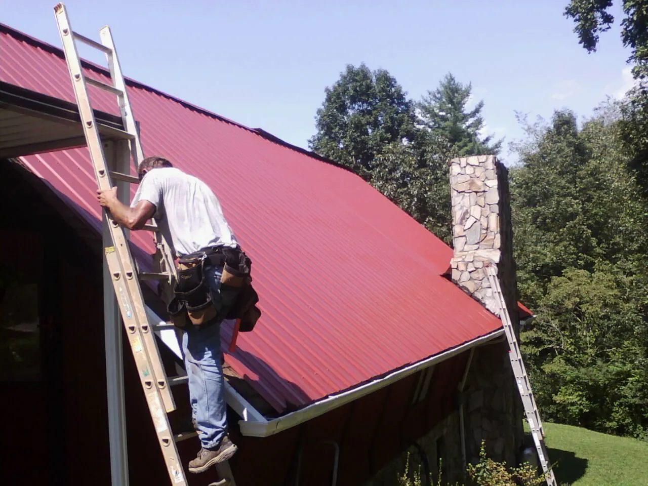 Roofer on a ladder beside a red metal roof and stone chimney — Soffit & Fascia Repair by Roofing Brothers in Elbe
